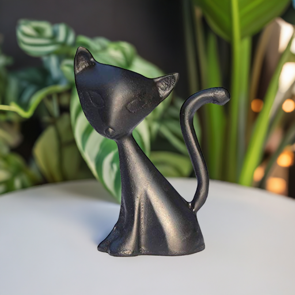 Matte black stylized cat ring holder with minimalist features and curled tail, sitting on a white surface with blurred green plants in the background.