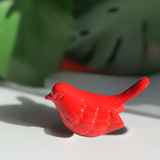 Small red bird figurine with detailed feather texture and raised tail casting shadow on white surface, green foliage blurred in background.