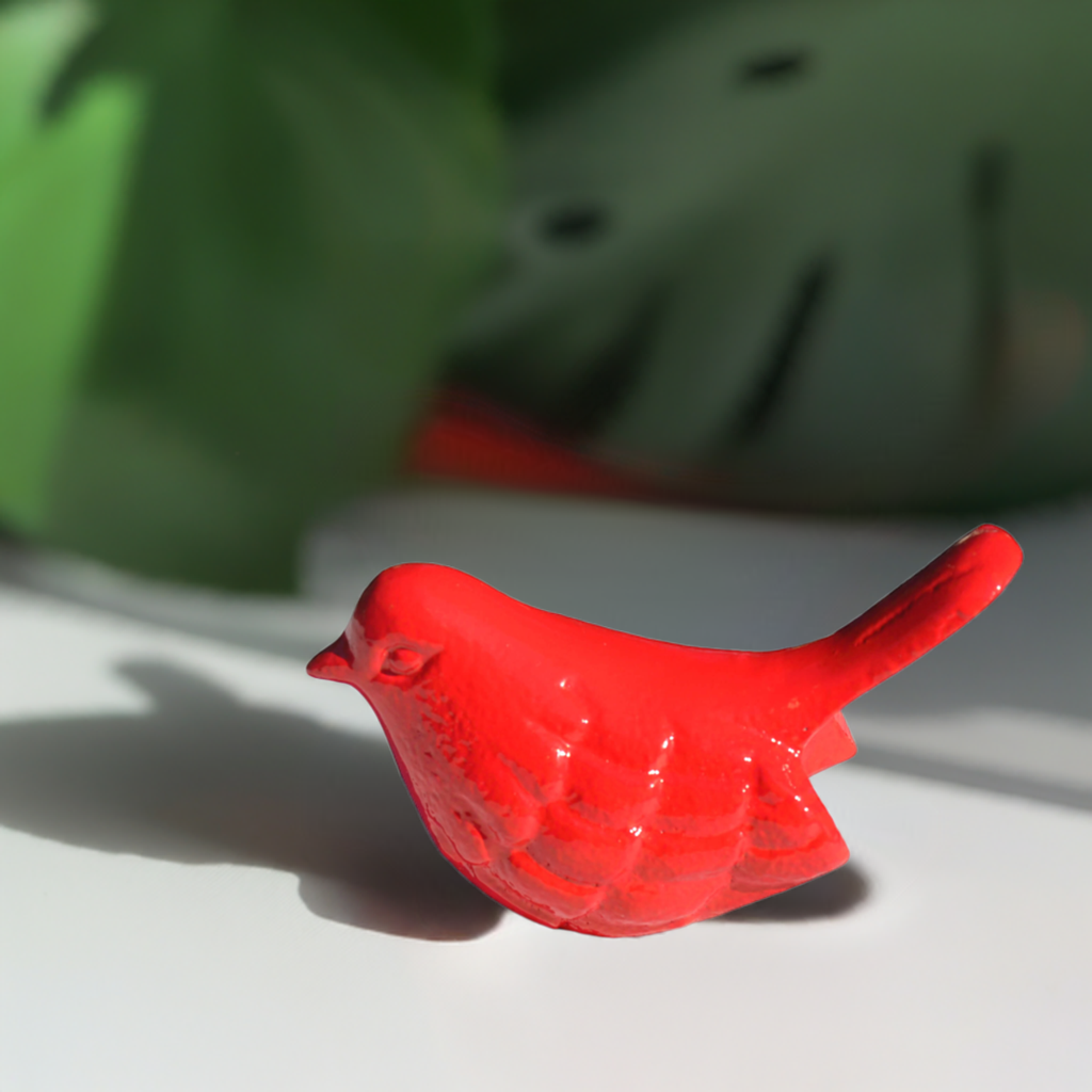 Small red bird figurine with detailed feather texture and raised tail casting shadow on white surface, green foliage blurred in background.
