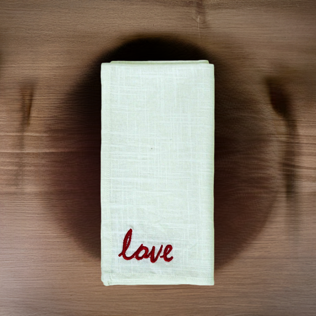 Cream-colored cloth napkin folded with red embroidered word ‘love’ in cursive on the corner, placed on a wooden surface with subtle grain.
