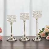 Set of three silver-toned aluminum candle holders with round crystal-studded square shades, arranged in ascending height on a wood surface with pink roses nearby.