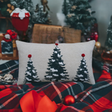 Rectangular cream-colored throw pillow featuring three stylized Christmas trees with snow and red pom-poms on top, placed on a red and dark plaid blanket with holiday decor in the background.