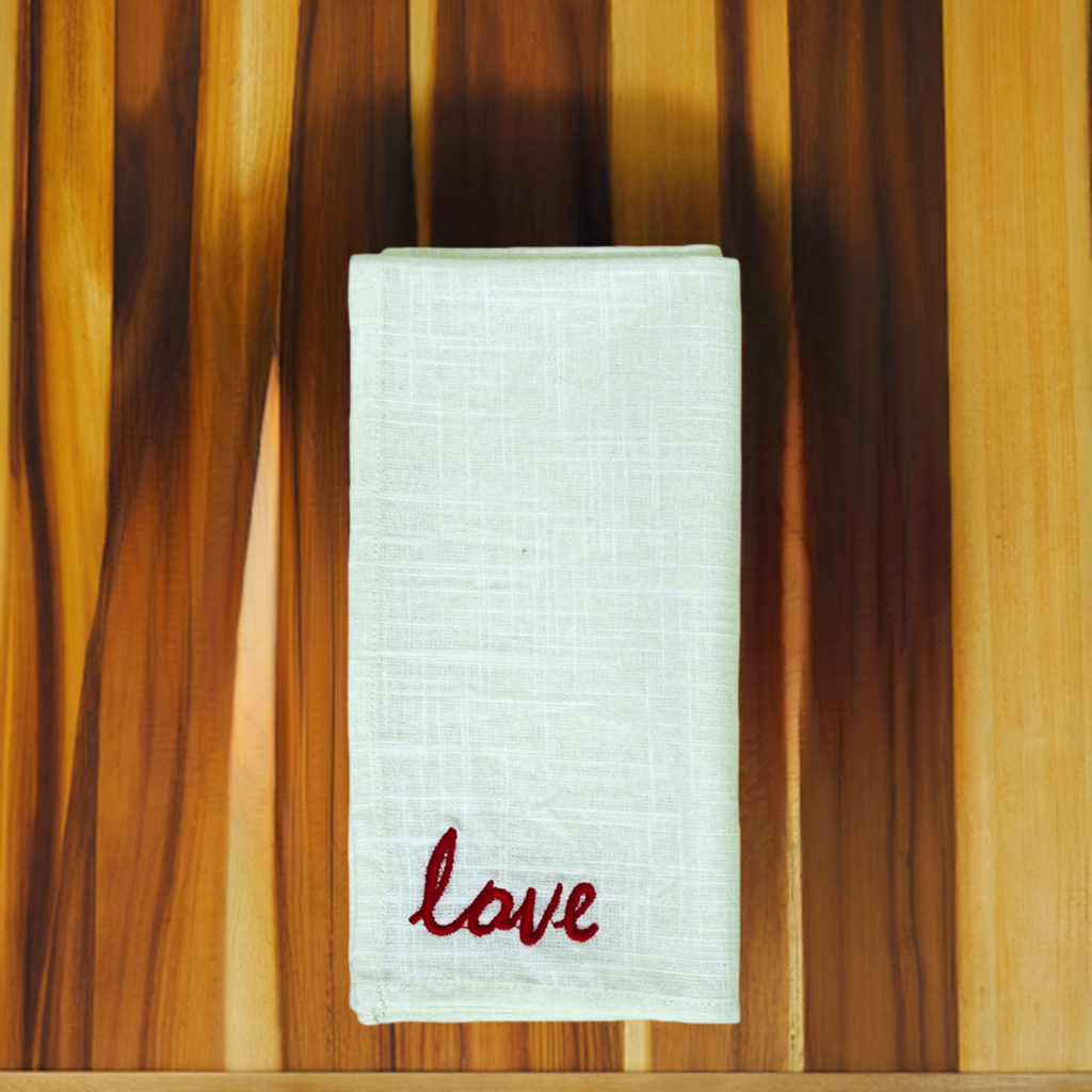 White cloth napkin folded neatly with red embroidery spelling 'love' at the bottom corner, placed on a wooden surface with varied brown tones.