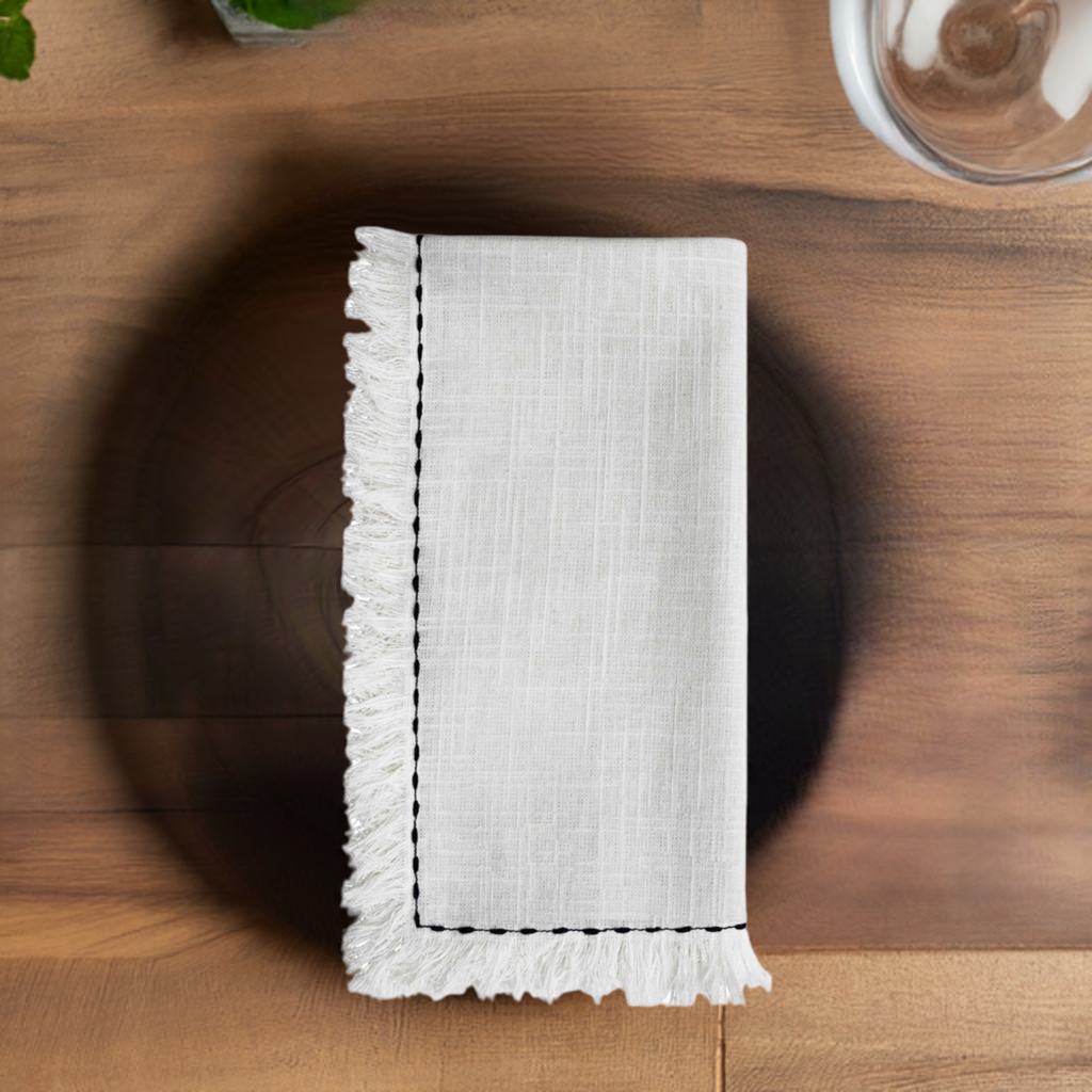 White linen napkin with black embroidered border and fringed edge, neatly folded on a round brown wooden plate set on a wooden tabletop.