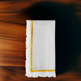 White linen napkin with yellow embroidered border and subtle texture, folded neatly on a polished dark wood surface with warm grain patterns.