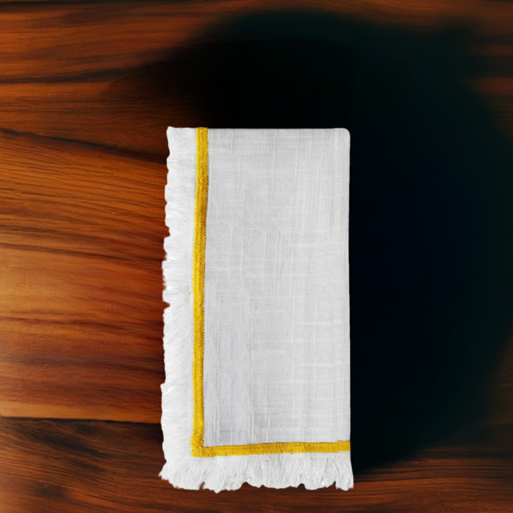 White linen napkin with yellow embroidered border and subtle texture, folded neatly on a polished dark wood surface with warm grain patterns.