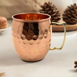 Copper-colored hammered metal mug with a gold-tone handle, shown on a light surface with blurred pine cones in the background.