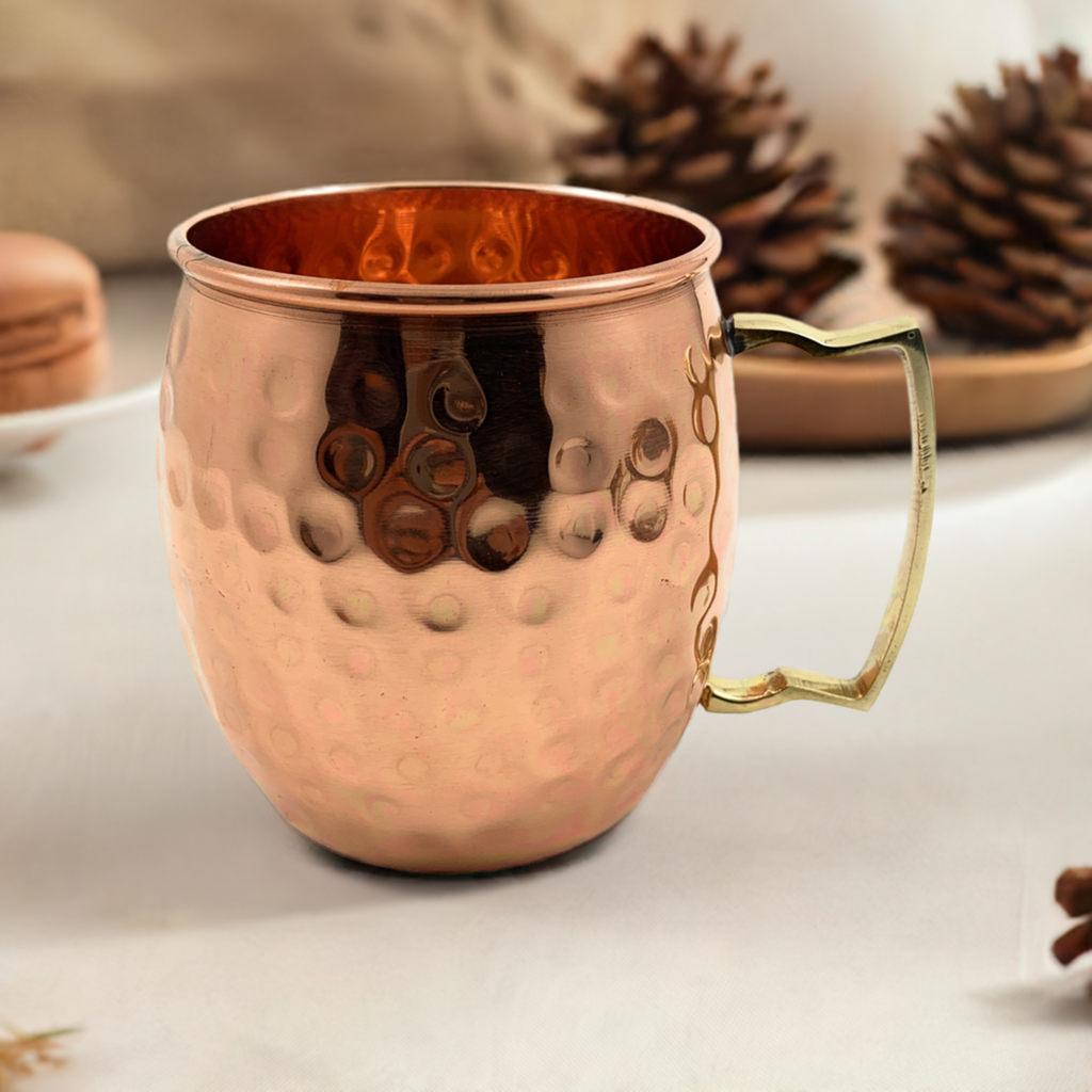 Copper-colored hammered metal mug with a gold-tone handle, shown on a light surface with blurred pine cones in the background.