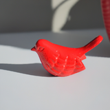 Small red bird figurine with textured wing and tail details, casting a distinct shadow on a white surface under natural light.