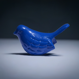 Small glossy solid blue bird figurine with carved feather details standing on a flat gray surface against a dark gradient background