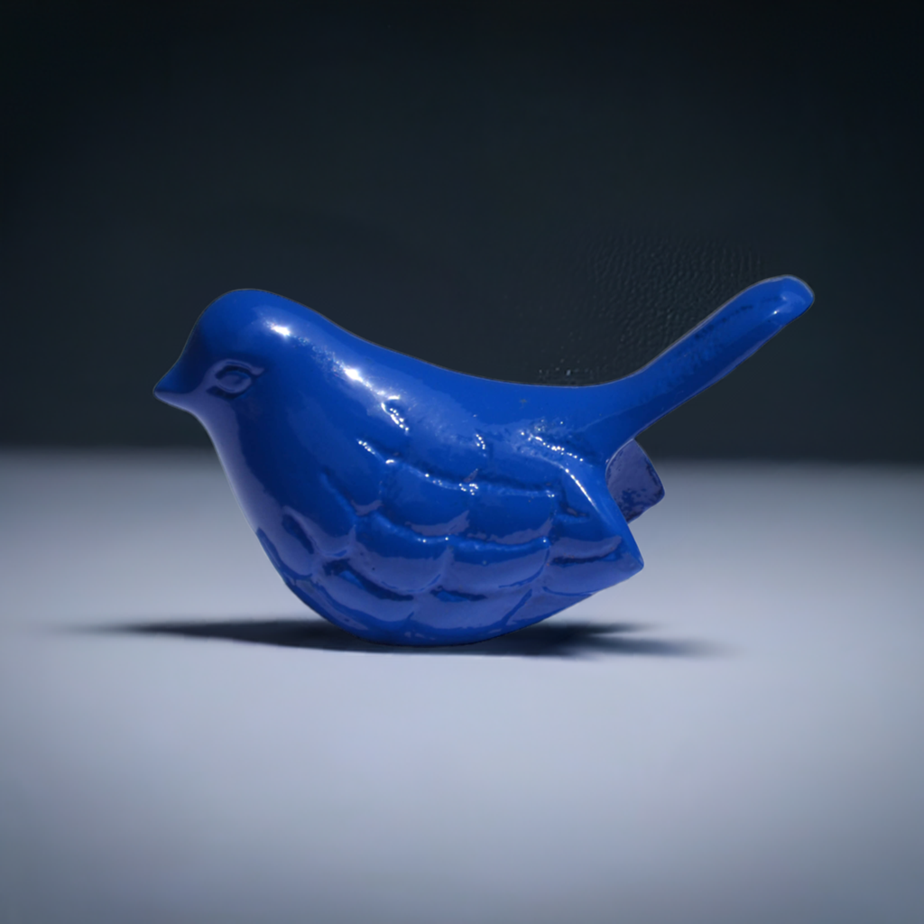 Small glossy solid blue bird figurine with carved feather details standing on a flat gray surface against a dark gradient background