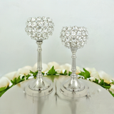 Two crystal ball-topped silver candle holders of different heights on a reflective surface surrounded by white flowers and green leaves.