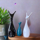 Three sleek ceramic deer figures with elongated necks in black, teal, and white matte finish, arranged on a wooden surface against a muted gray backdrop.