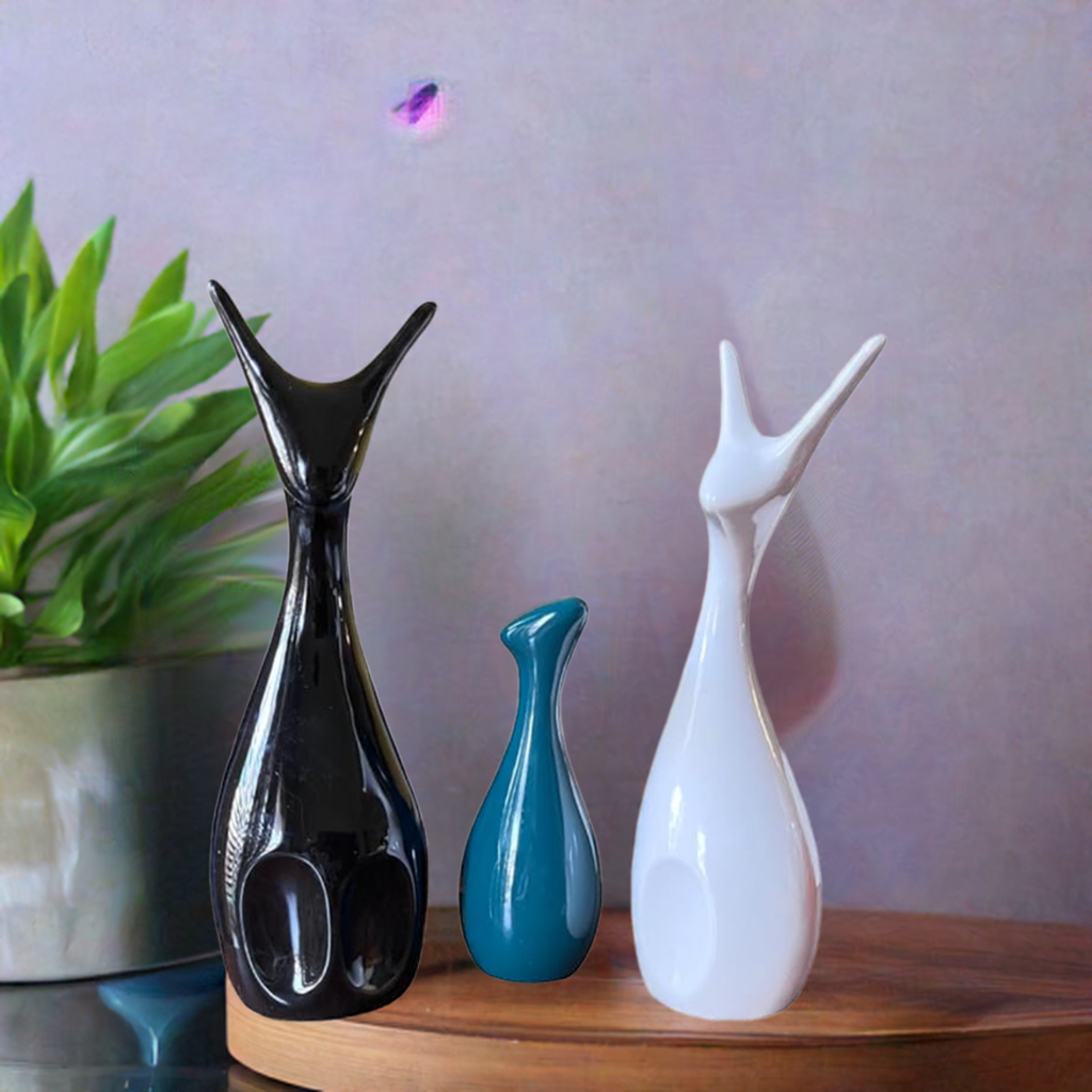 Three sleek ceramic deer figures with elongated necks in black, teal, and white matte finish, arranged on a wooden surface against a muted gray backdrop.