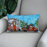 Rectangular pillow on a beige armchair featuring an illustration of an orange pickup truck towing a decorated trailer with Christmas trees under a blue sky.