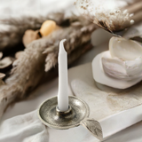 Silver tapered candlestick holder dish with a decorative leaf accent, holding an unlit white taper candle, surrounded by soft neutral dried plants and fabric.