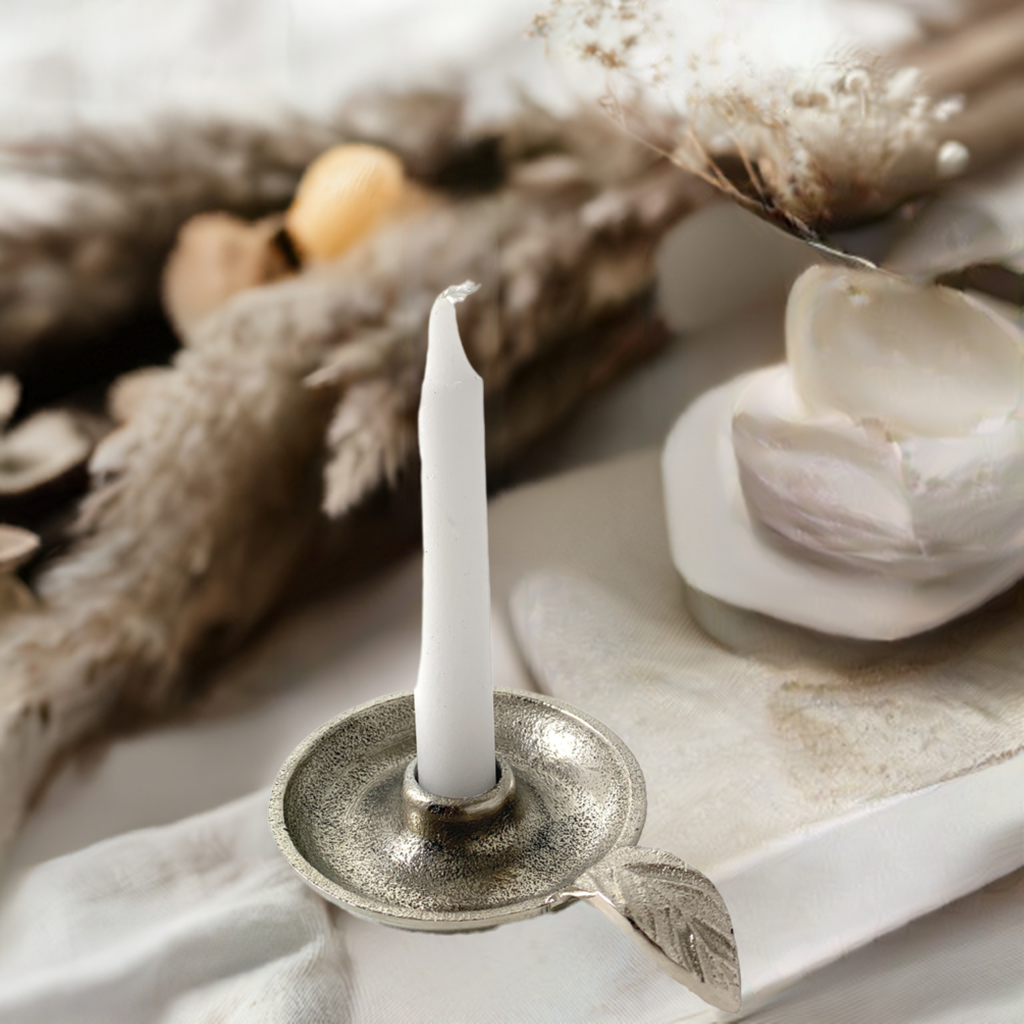 Silver tapered candlestick holder dish with a decorative leaf accent, holding an unlit white taper candle, surrounded by soft neutral dried plants and fabric.
