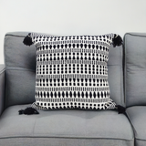 Square black and white throw pillow with geometric pattern and tassels on each corner, placed centered on a gray couch cushion.