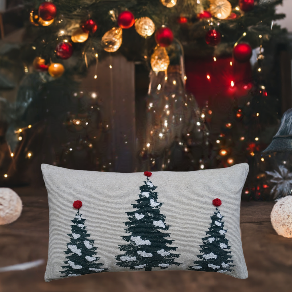 Rectangular beige pillow with three green Christmas trees, each topped with a red pom-pom, placed in front of a decorated, softly lit Christmas tree background.