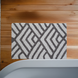 Rectangular bath rug with a grey and ivory geometric diamond pattern placed on honey-toned wooden floor next to a curved light gray bathtub edge.