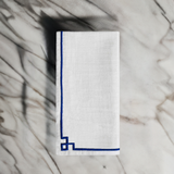 White linen napkin folded on marble surface with simple dark blue embroidered border, featuring corner geometric detail.