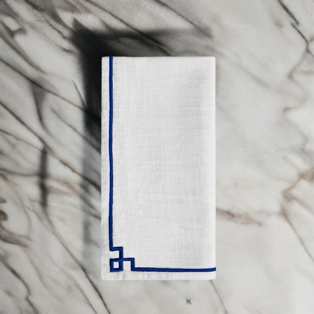 White linen napkin folded on marble surface with simple dark blue embroidered border, featuring corner geometric detail.