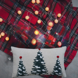Rectangular beige throw pillow with three dark green Christmas trees topped with red pom-poms, set against a blurred background of red plaid and warm string lights.