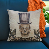 Square throw pillow with embroidered skull wearing a tall top hat, flanked by two birds and ornate decorative swirls on beige fabric.
