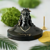 Black resin bust of Adiyogi with detailed facial features, a snake around the neck, and 'Adiyogi' inscribed on the base, set against a blurred white background with green leaves.