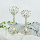 Set of two silver crystal candle holders with round, gem-studded cups on tall, slender stems and circular stepped bases, placed on light fabric with a green vine.