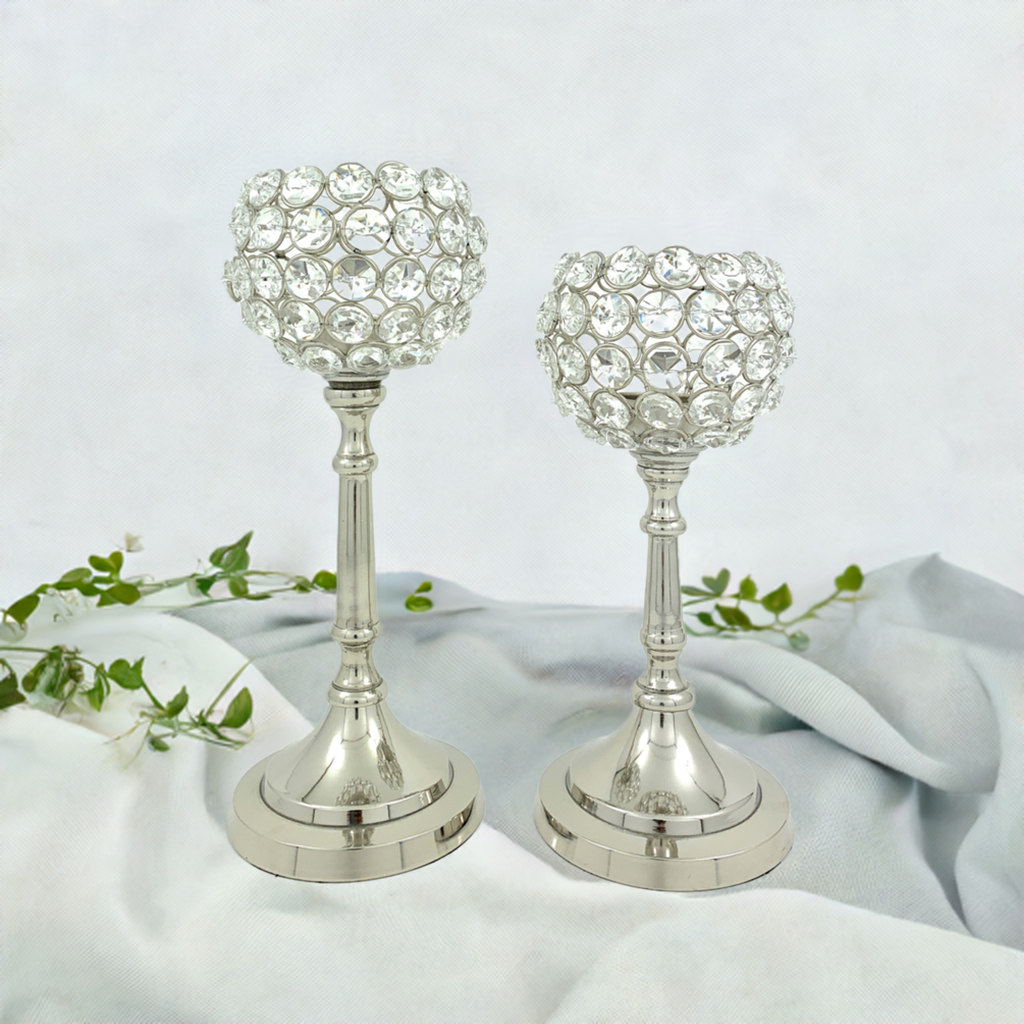 Set of two silver crystal candle holders with round, gem-studded cups on tall, slender stems and circular stepped bases, placed on light fabric with a green vine.