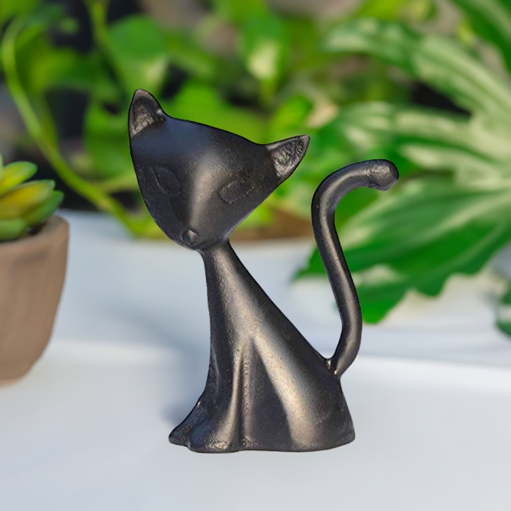 Small black matte cat figurine with minimalist features and a curled tail, set against a blurred background with green leaves and small potted plants.