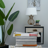 Decorative couch pillow with gray and white stripes, accented by mustard yellow, rust, and pink embroidery braids, featuring tassels on all four corners