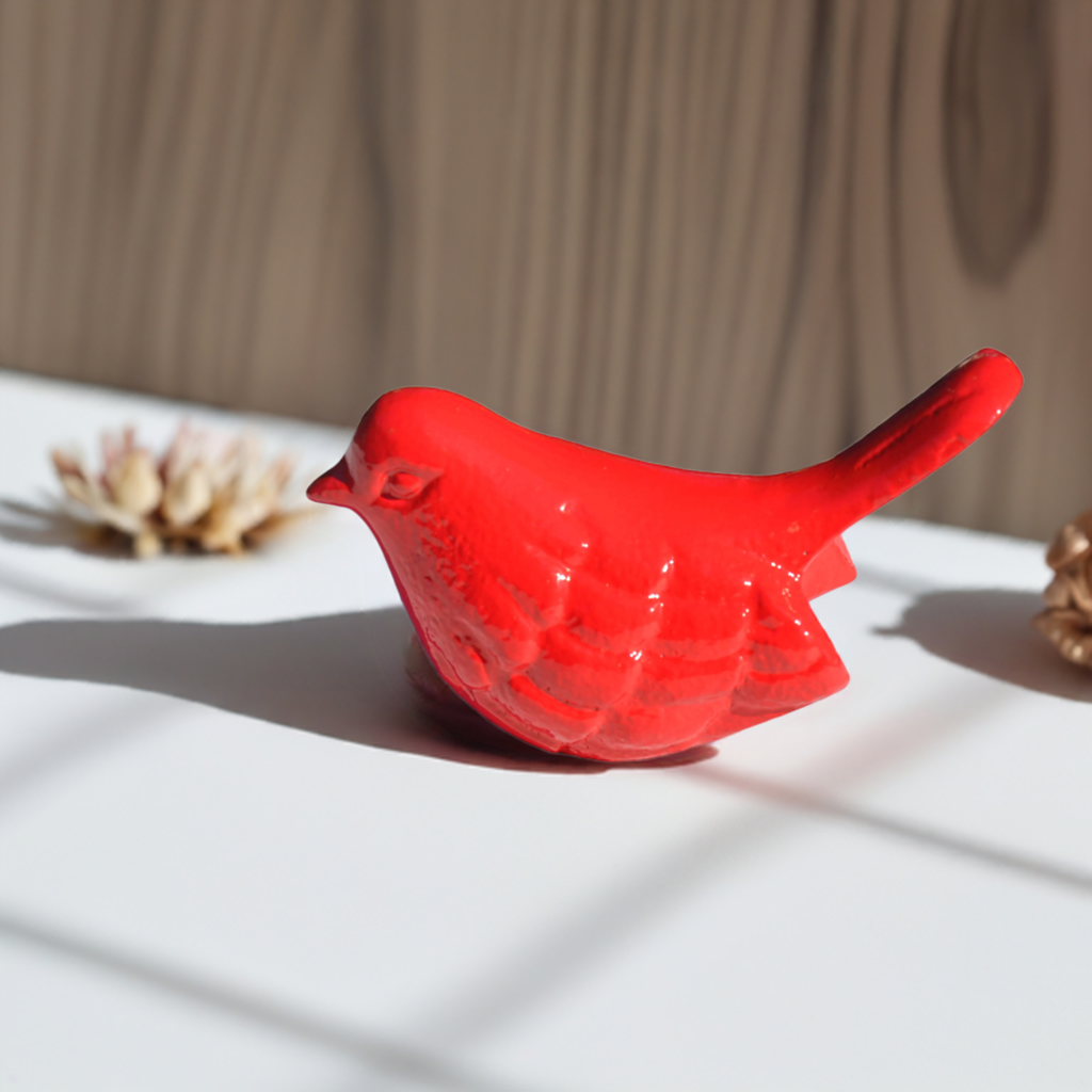 Small red glossy bird figurine with embossed wing details casting a shadow on a white surface, with blurred natural wooden background and light patterns.