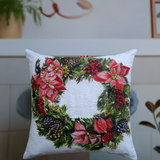 Square white throw pillow featuring a festive Christmas wreath design with red poinsettias, pinecones, and green foliage on the front center.