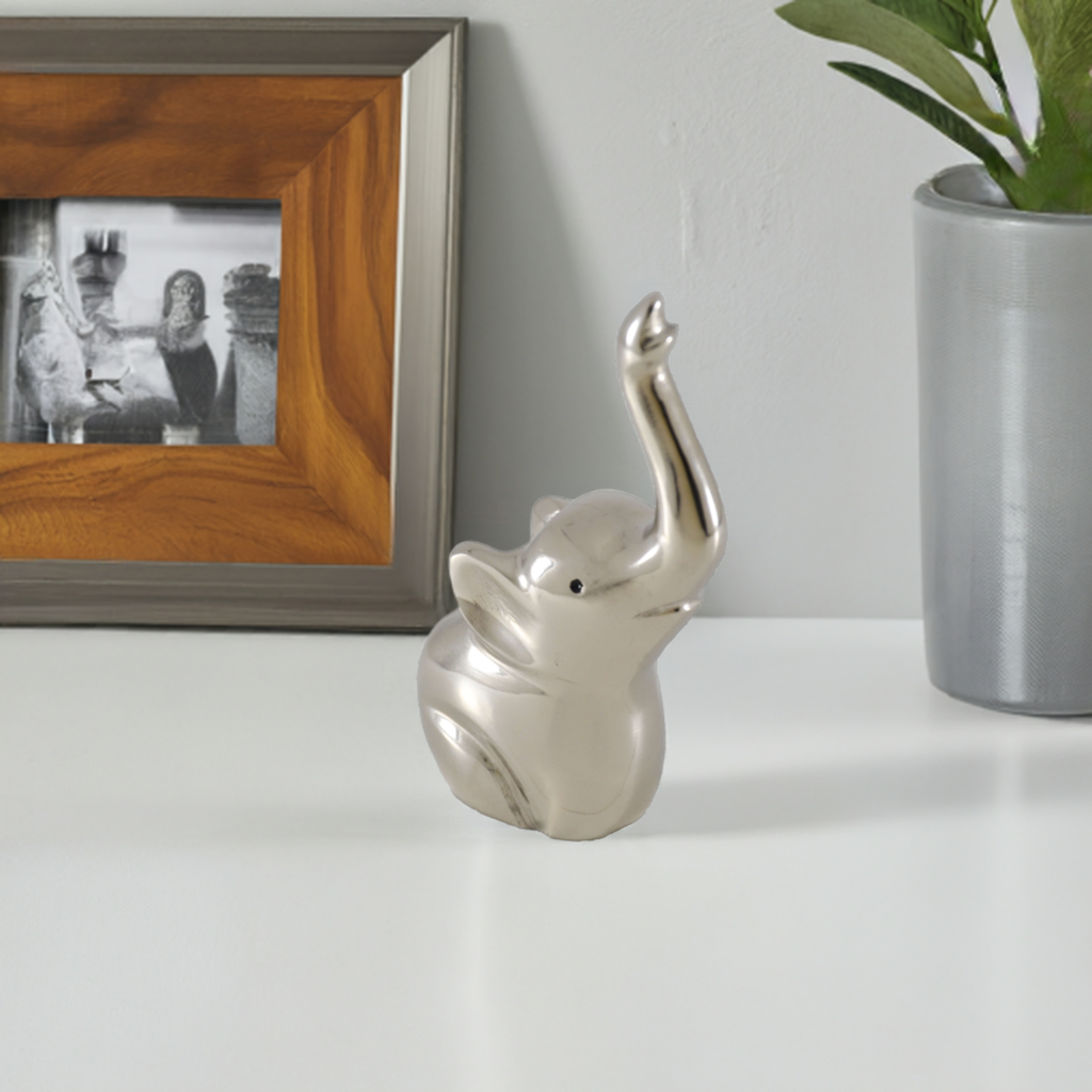 Small silver-finished elephant-shaped ring holder with raised trunk, sitting on a white surface between a framed photo with wood and gray border and a gray cylindrical pot with green plant.