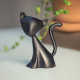 Black cat figurine with an elongated body and raised curved tail, sitting on a white surface with a blurred green plant and brown pot in the background.