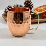 Single hammered copper mug with a shiny brass handle, shown against a backdrop of pine cones and dried citrus slices on a wooden tray.