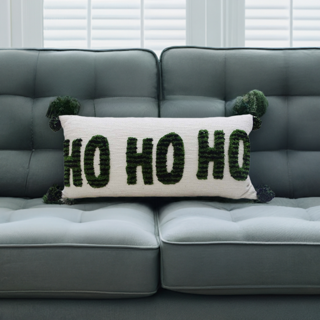 Rectangular white pillow with green embroidered 'HO HO HO' text and green pom-poms at each corner, placed on a gray tufted couch against white window blinds.