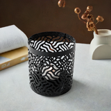 Black cylindrical votive candle holder with intricate wavy cutout patterns allowing candlelight to shine through, placed on a gray surface near a book and a minimalist white vase.