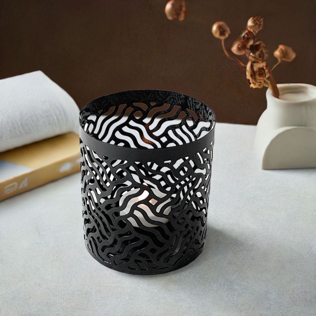 Black cylindrical votive candle holder with intricate wavy cutout patterns allowing candlelight to shine through, placed on a gray surface near a book and a minimalist white vase.