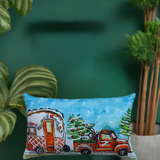 Throw pillow with illustration of vintage orange truck carrying decorated Christmas trees and a trailer against a blue sky background, surrounded by green plants.
