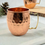 Copper hammered mug with a brass handle on a white cloth, showing its textured surface and rounded shape with a matching mug blurred in the background