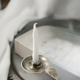 Silver-toned metal taper candle holder with a textured dish and a small leaf-shaped handle, holding an unlit white candle, on a blurred fabric and box background.