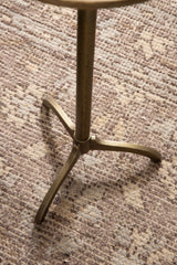 Close-up of the tripod base of a round metal side table with a muted brass finish, set against a textured beige patterned rug.