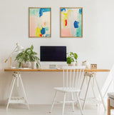 Two framed abstract paintings featuring blue, yellow, red, and pink brushstrokes hang above a minimalist wooden desk with white sawhorse legs, holding a computer, plants, and a white chair.