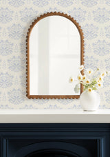 Arched wall mirror with scalloped wooden frame hangs above white shelf holding round white vase with pale yellow flowers against blue floral wallpaper.