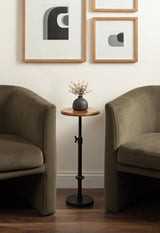 Small round wooden adjustable-height table with black metal base between two olive green velvet armchairs, holding a black vase with dried flowers, beneath framed abstract art.