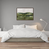Framed canvas painting of green rolling hills with broad brushstrokes in various green shades, mounted on a gray wall above a white bed with white bedding and a white bedside lamp.