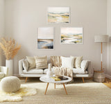 Living room with neutral beige tones featuring a light gray sofa with patterned cushions, three framed abstract landscape canvases above, a round marble coffee table, and dried pampas in a white vase.